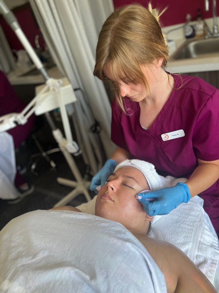 This is an image with a female student in blue gloves performing an esthetics procedure on a client lying on a table wearing a white covering.